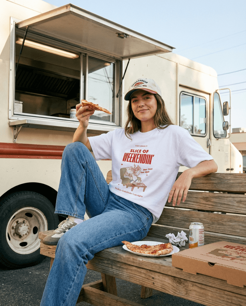 Woman enjoying pizza while wearing an Oversized Pizza T-Shirt – Slice of Weekendin’ (White) in front of a food truck.