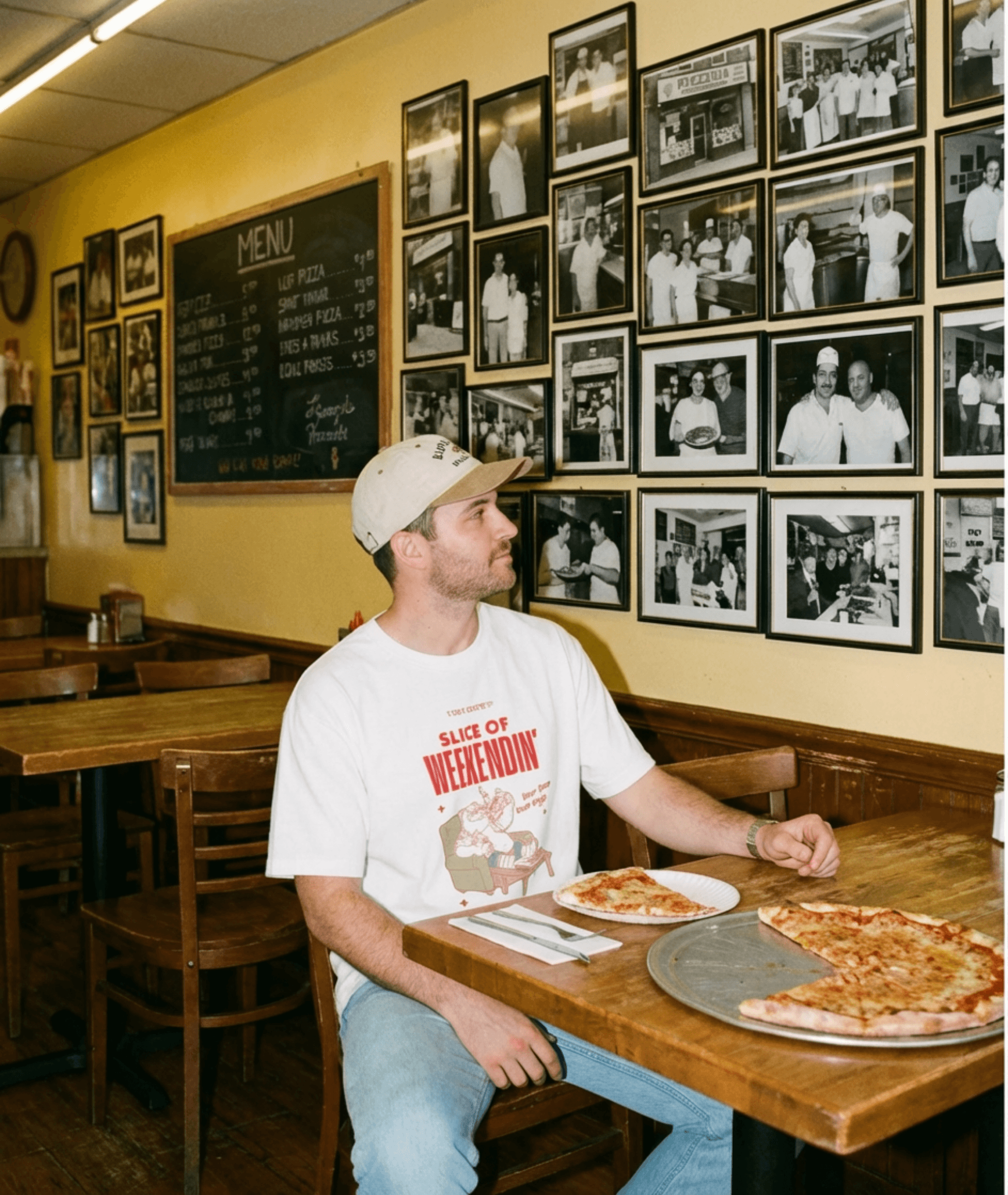 A man wearing an Oversized Pizza T-Shirt – Slice of Weekendin’ (White) sits at a diner table with pizza.