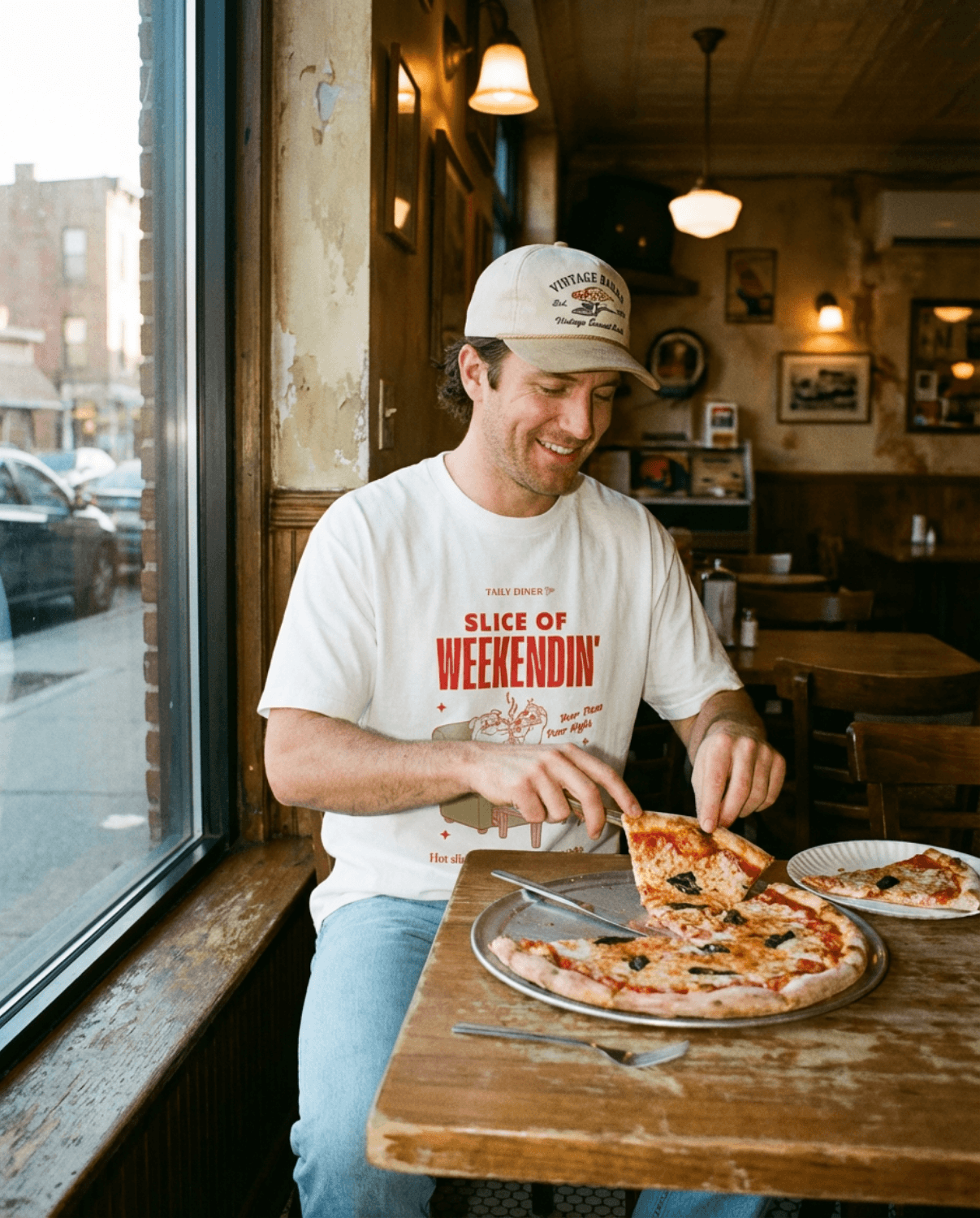 Man enjoying pizza while wearing an Oversized Pizza T-Shirt – Slice of Weekendin’ (White) in a diner setting.