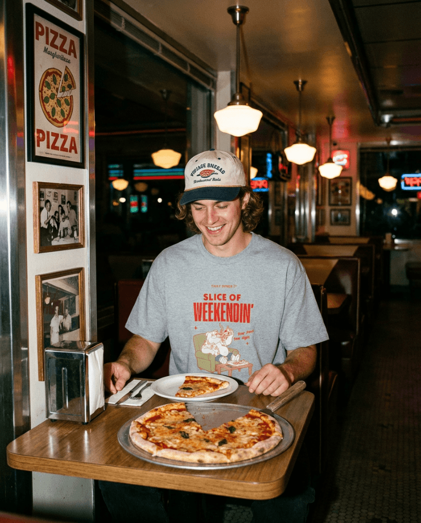 A person wearing an Oversized Pizza T-Shirt – Slice of Weekendin’ (Athletic Heather) enjoys pizza at a diner.