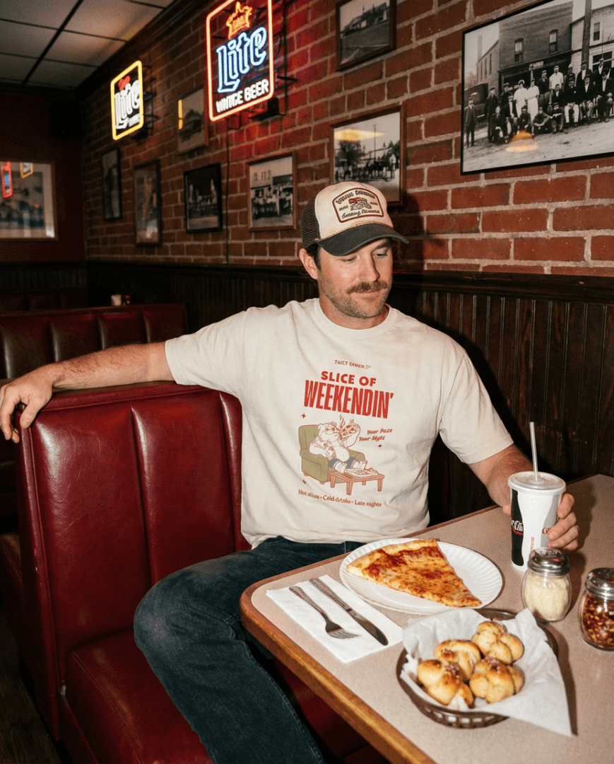 Man wearing Oversized Pizza T-Shirt – Slice of Weekendin’ (Ecru) in diner with pizza and drink.
