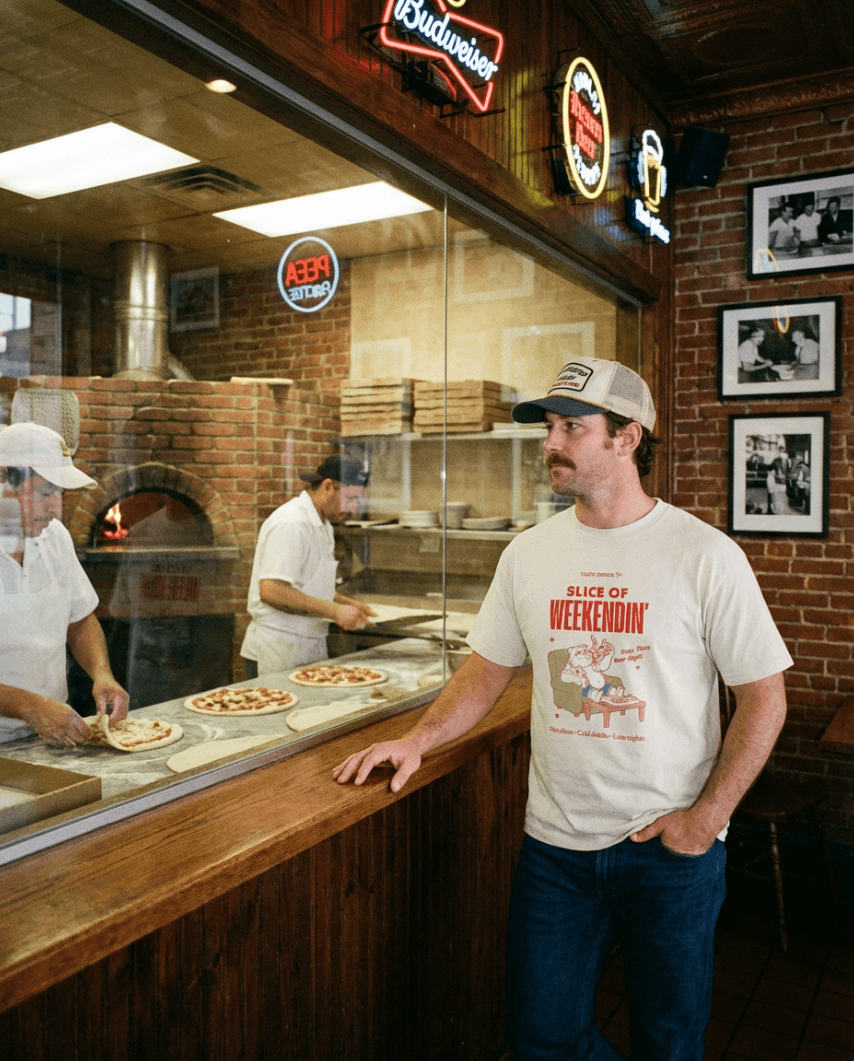A man wearing an Oversized Pizza T-Shirt – Slice of Weekendin’ (Ecru) at a pizzeria.