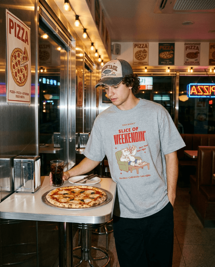 Person wearing Oversized Pizza T-Shirt – Slice of Weekendin’ (Athletic Heather) in a diner with pizza.