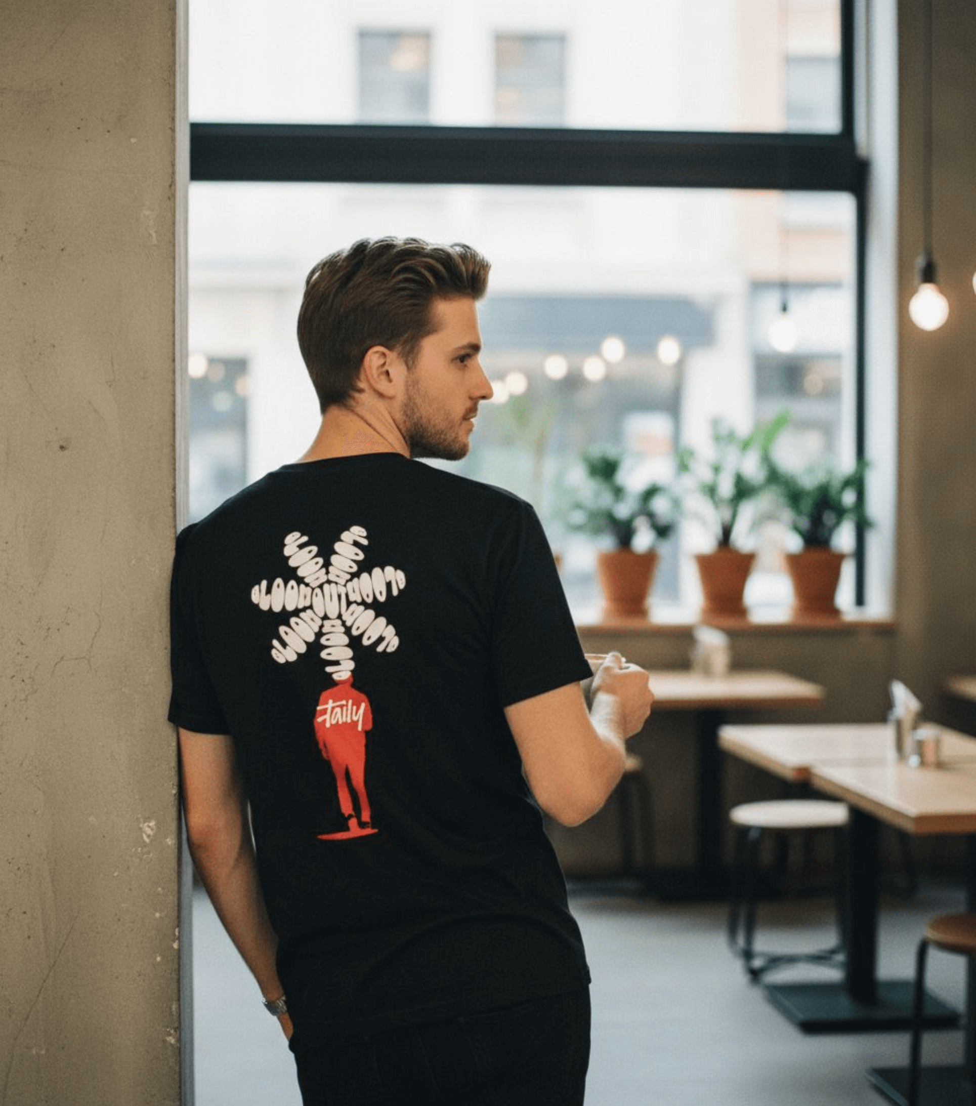 Man wearing Taily Bloom streetwear graphic t-shirt in black with bold back design in urban cafe setting.