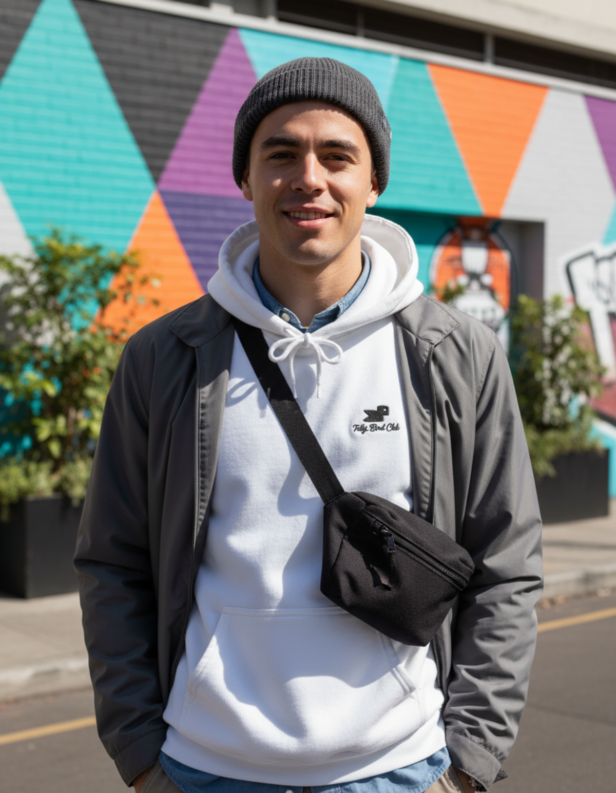 Young man wearing Taily Bird embroidered hoodie and black bag in front of colorful mural.