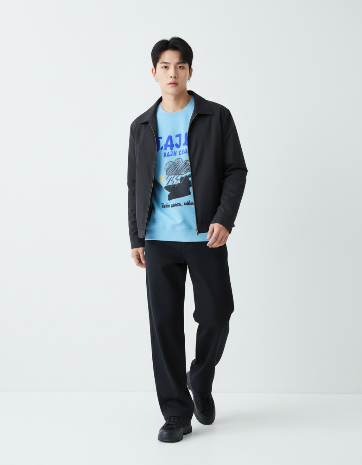 Model wearing Taily Rain Club sweatshirt in Sky with stylish black jacket and pants standing against a minimalist background.