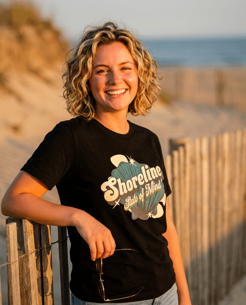 Shoreline State of Mind Tee – Black worn by a woman at the beach, featuring a vintage seashell graphic.
