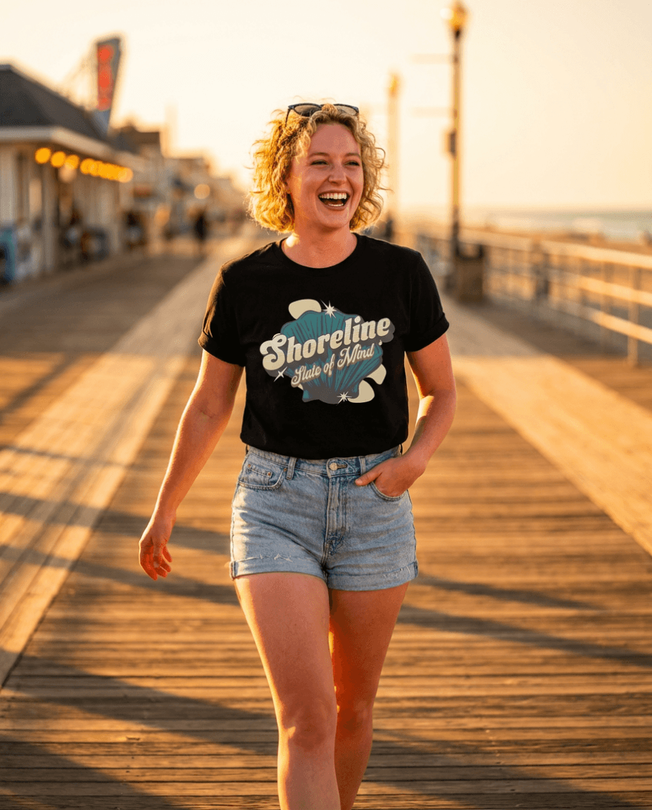 Woman walking on boardwalk wearing Shoreline State of Mind Tee – Black with seashell graphic at sunset.
