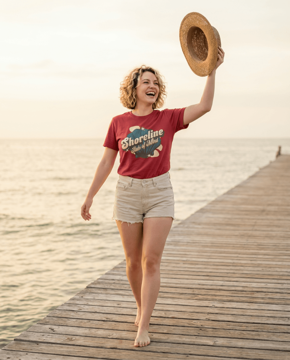 Woman wearing the Shoreline State of Mind Tee – Crimson, walking on a pier and holding a straw hat.