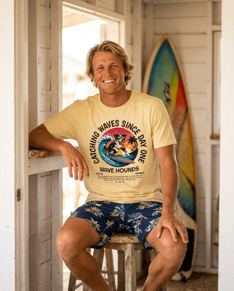Man wearing Wave Hounds Surf Tee – Banana, sitting on a stool with surfboards in the background.