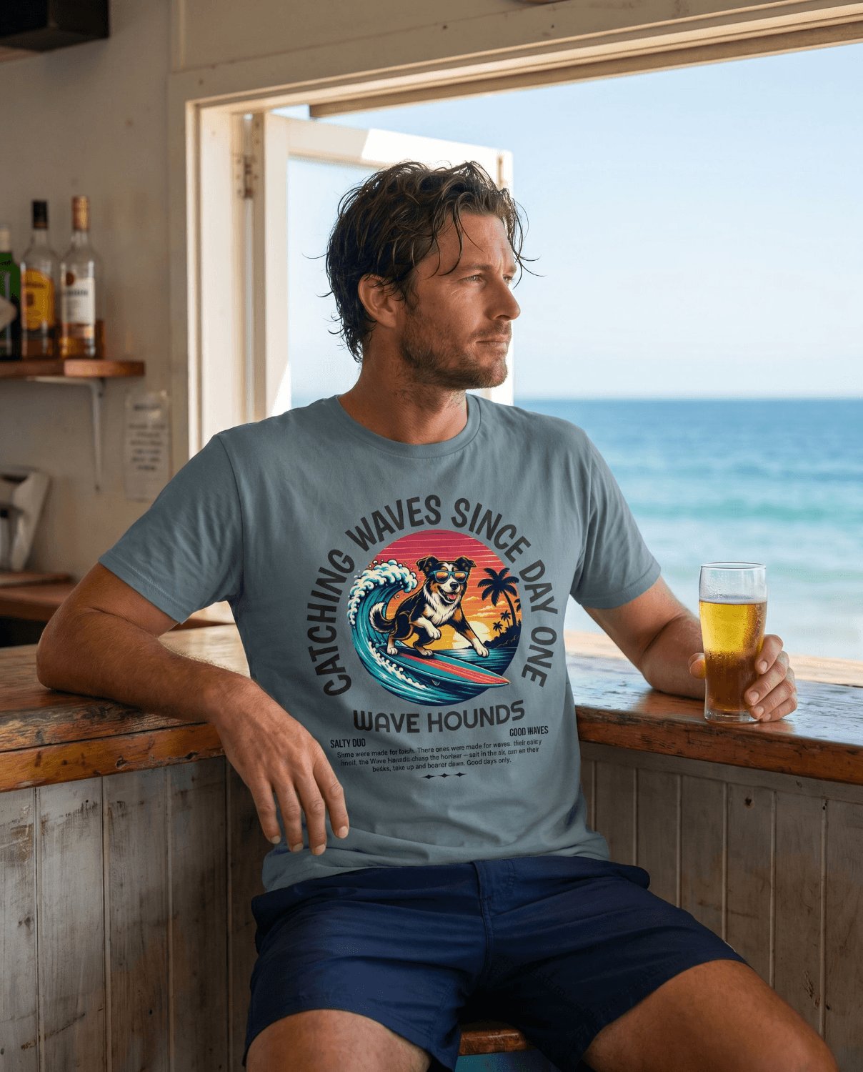 Man wearing Wave Hounds Surf Tee – Ice Blue, enjoying a drink with ocean view, retro surf graphic on shirt.