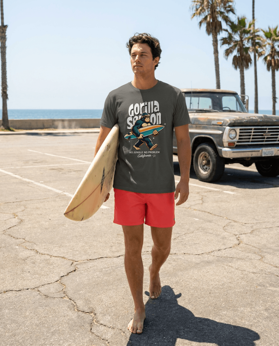 Gorilla Season Surf Tee – Pepper worn by a man walking on a beach, carrying a surfboard with palm trees in the background.