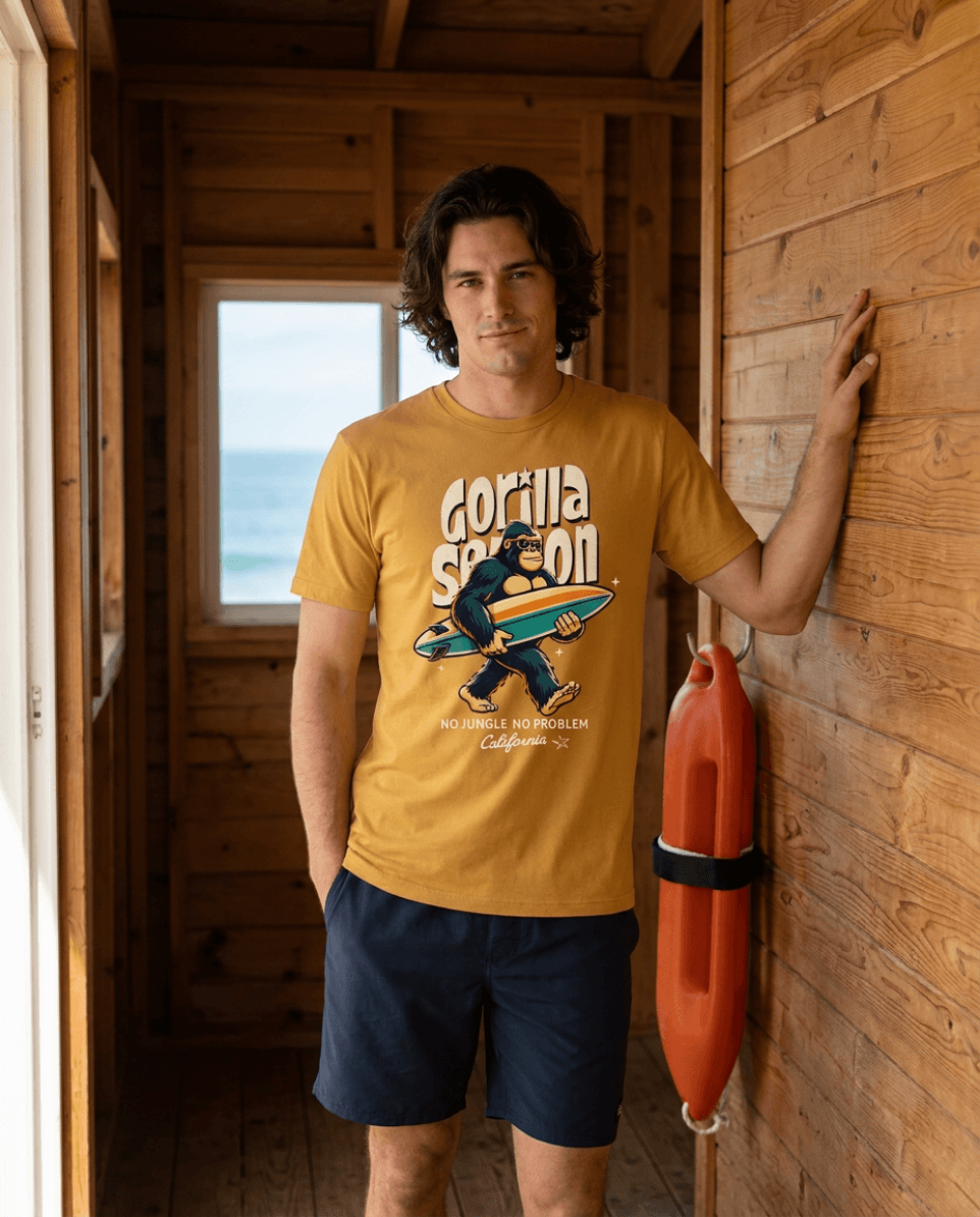 Model wearing Gorilla Season Surf Tee – Mustard inside a beach shack with surfboard.