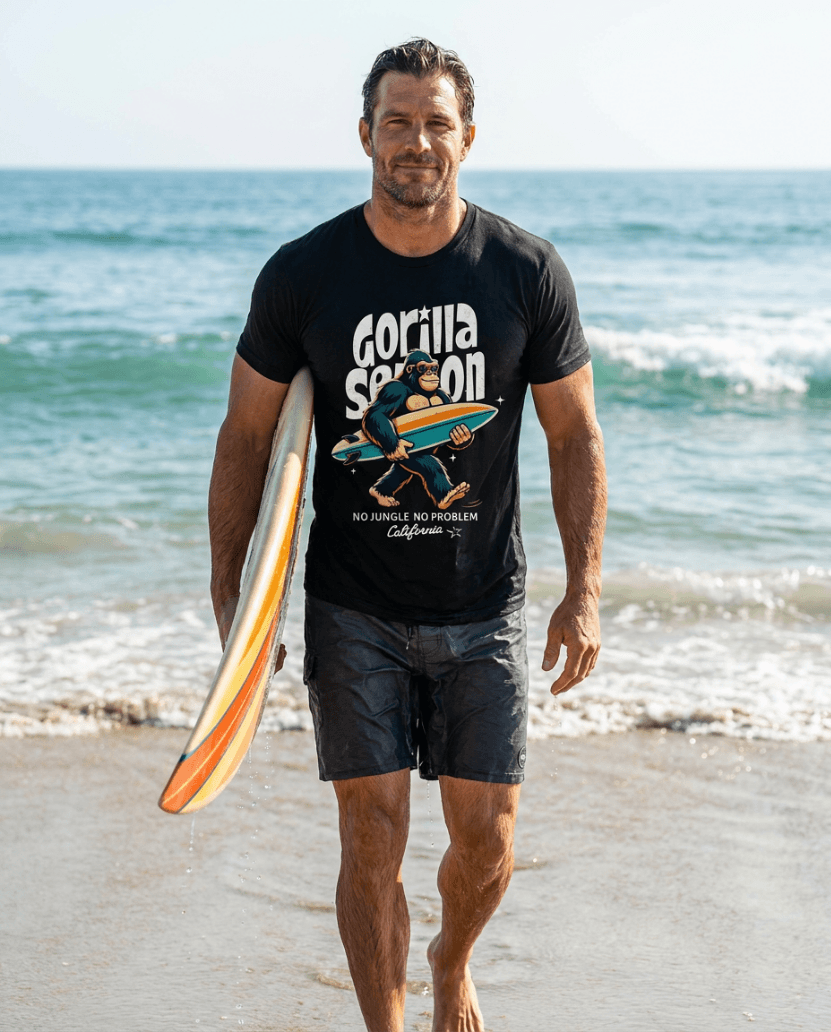 Man wearing Gorilla Season Surf Tee – Black, walking on the beach with a surfboard and ocean in the background.