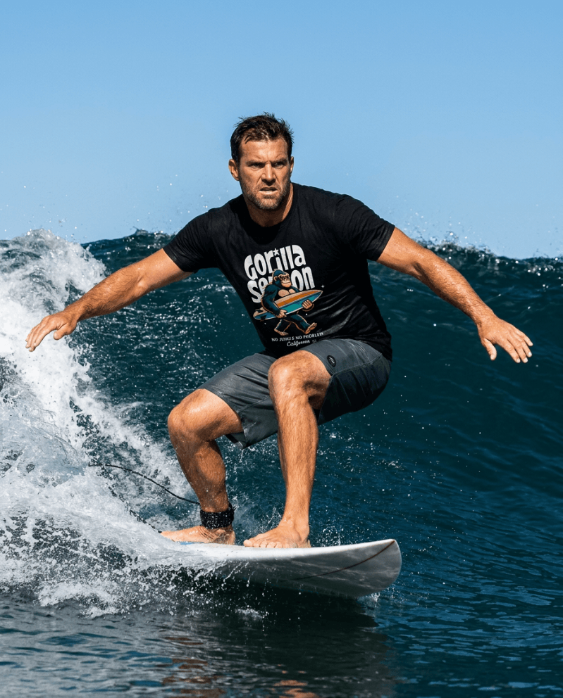 Man surfing in a wave wearing Gorilla Season Surf Tee – Black and shorts, showcasing a bold surf graphic design.