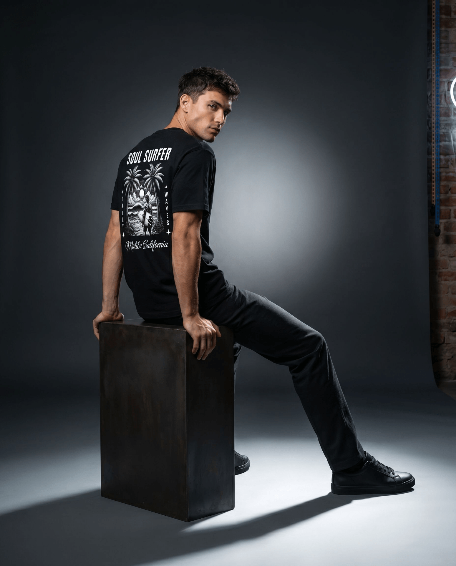 Model wearing Soul Surfer Malibu Tee with palm tree graphic, seated on a cube in a studio setting.