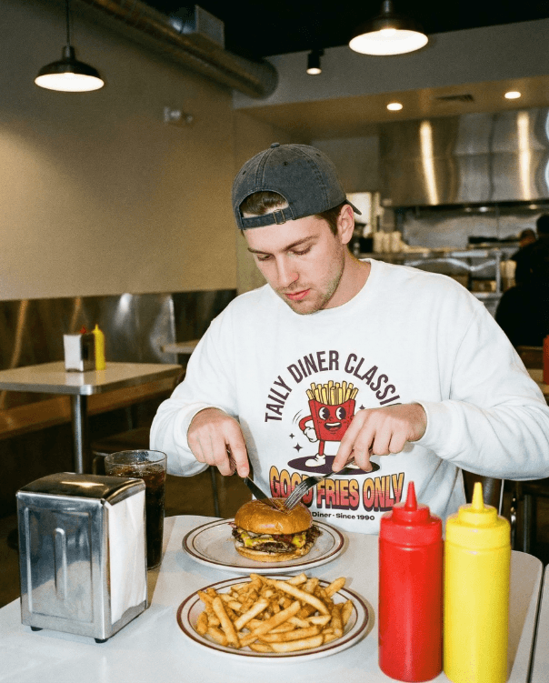 Man enjoying a meal wearing the Good Fries Only Sweatshirt – Taily Diner (White) in a retro diner setting.