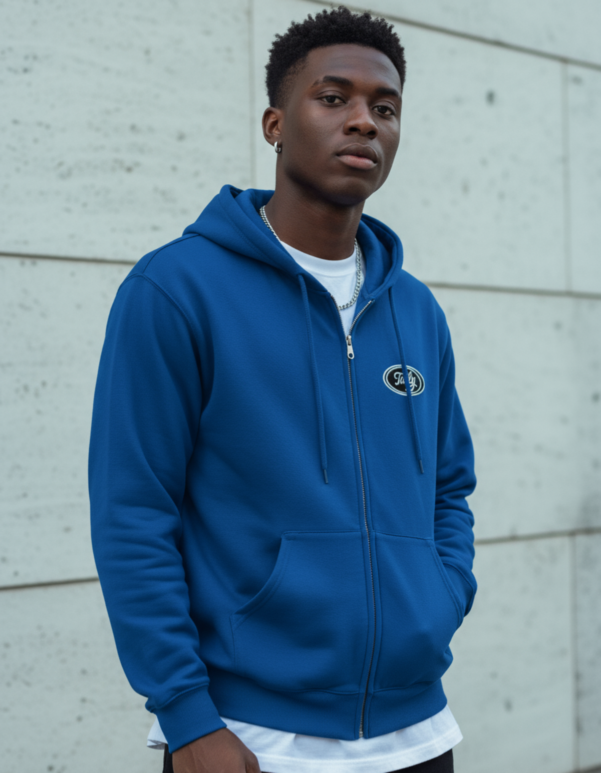 Model wearing a Taily Zip Hoodie in royal blue with embroidered logo against a grey concrete wall.