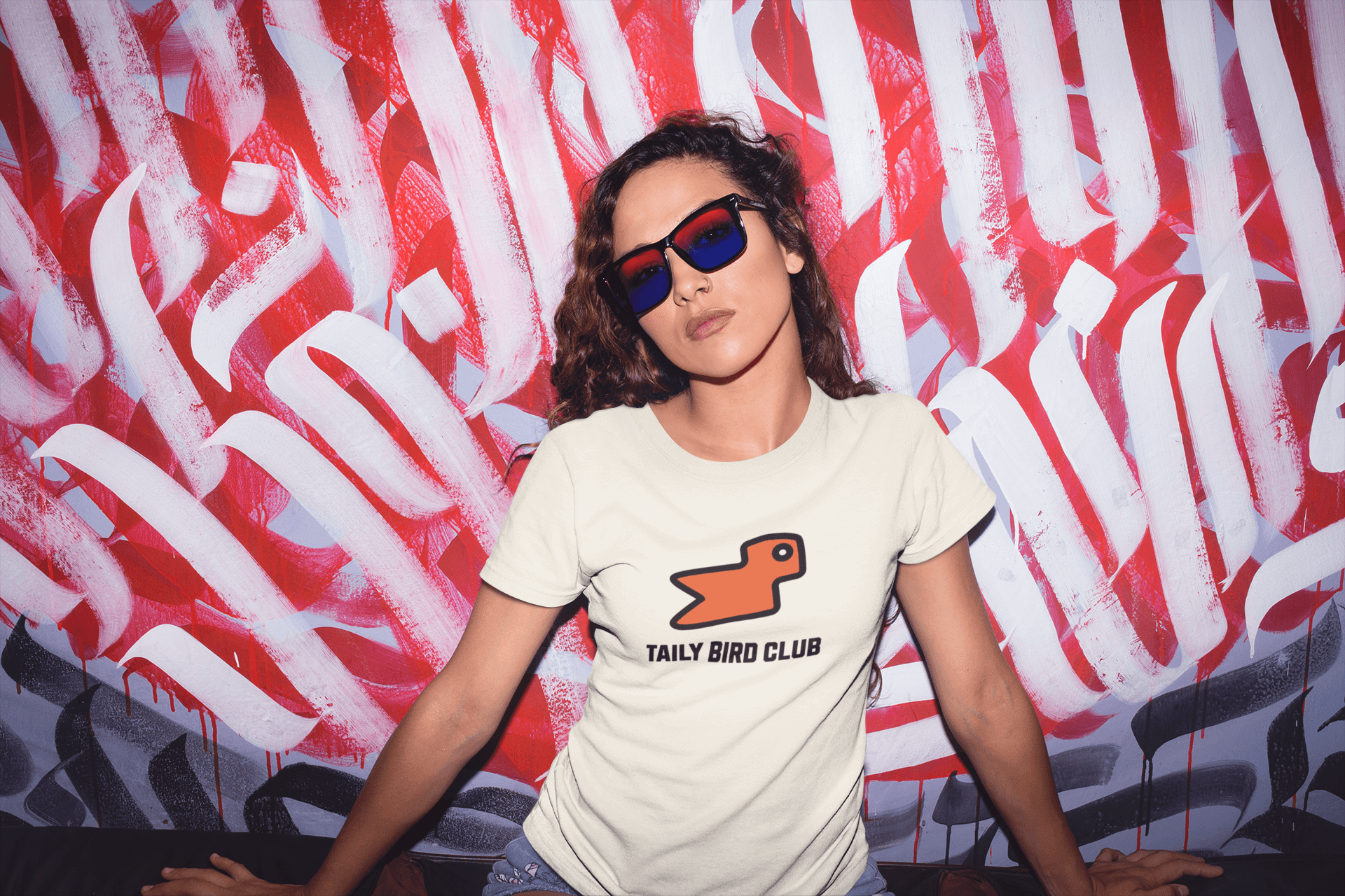 Model wearing Taily Bird Club boxy t-shirt with orange bird design against vibrant graffiti background.
