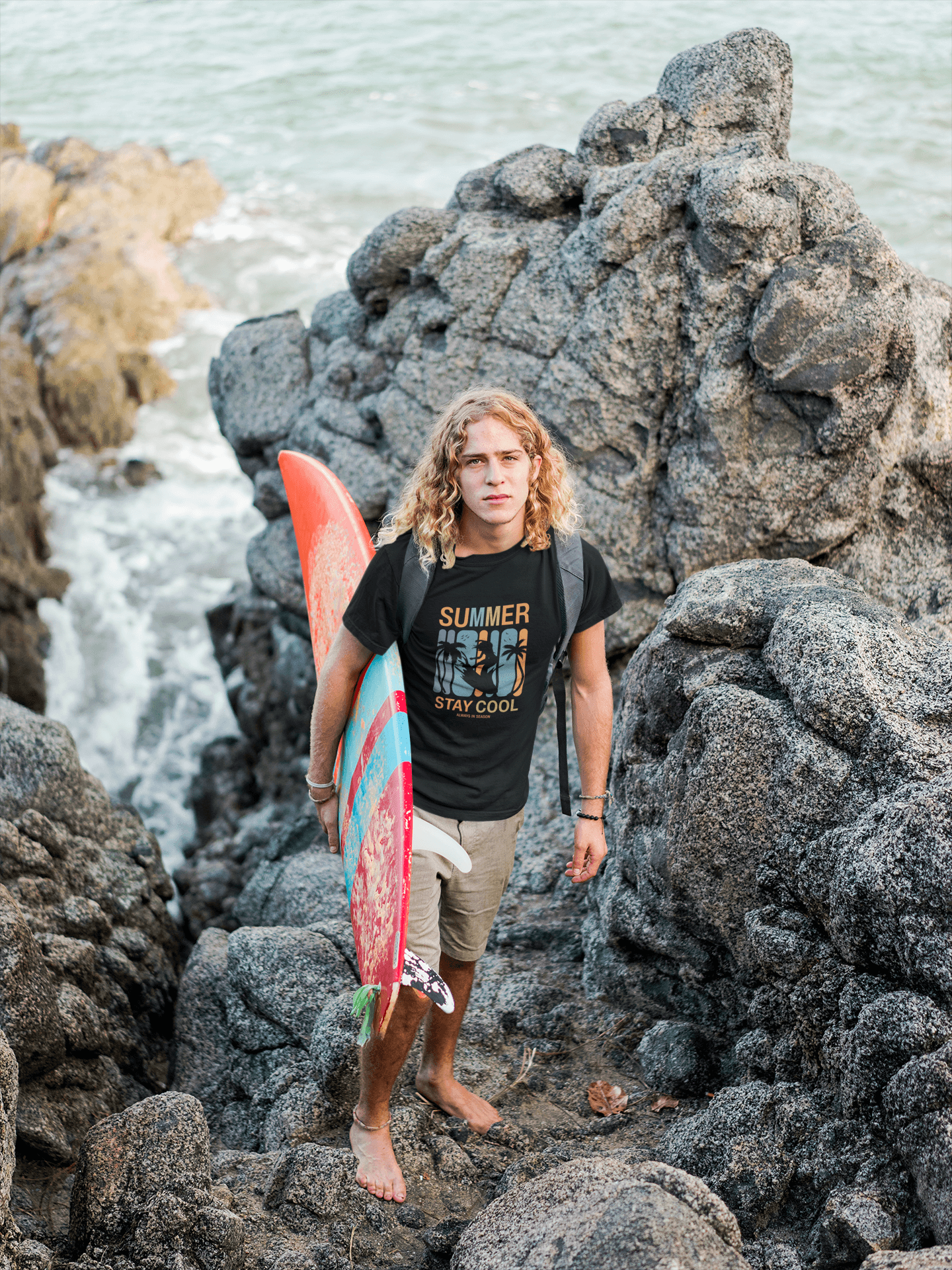 Man with long hair in a Summer Stay Cool T-shirt holding a surfboard, standing on rocky beach makes for a dynamic summer vibe.