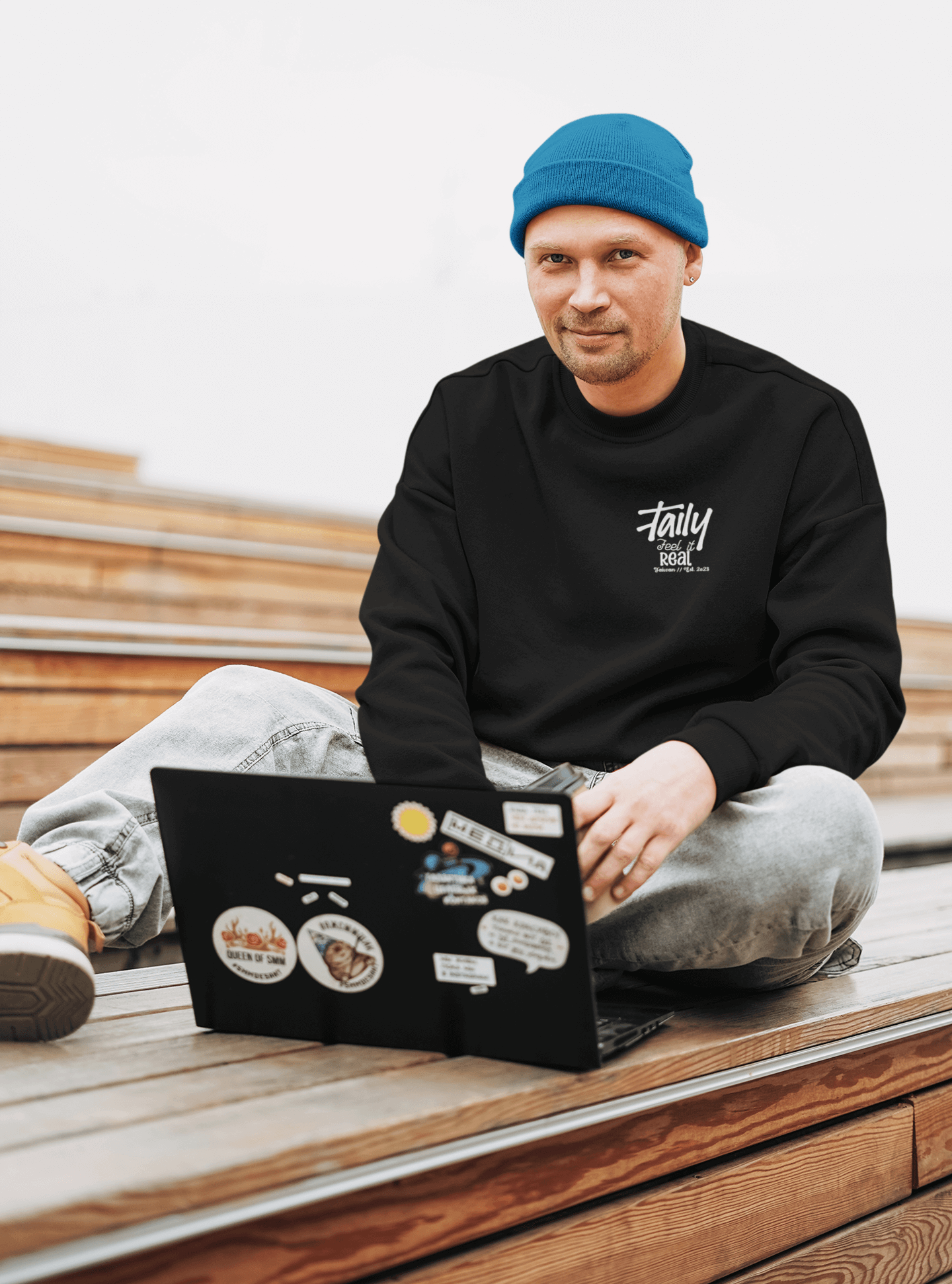 Man wearing Taily Feel It Real black sweatshirt with chest logo, sitting on steps with a laptop.