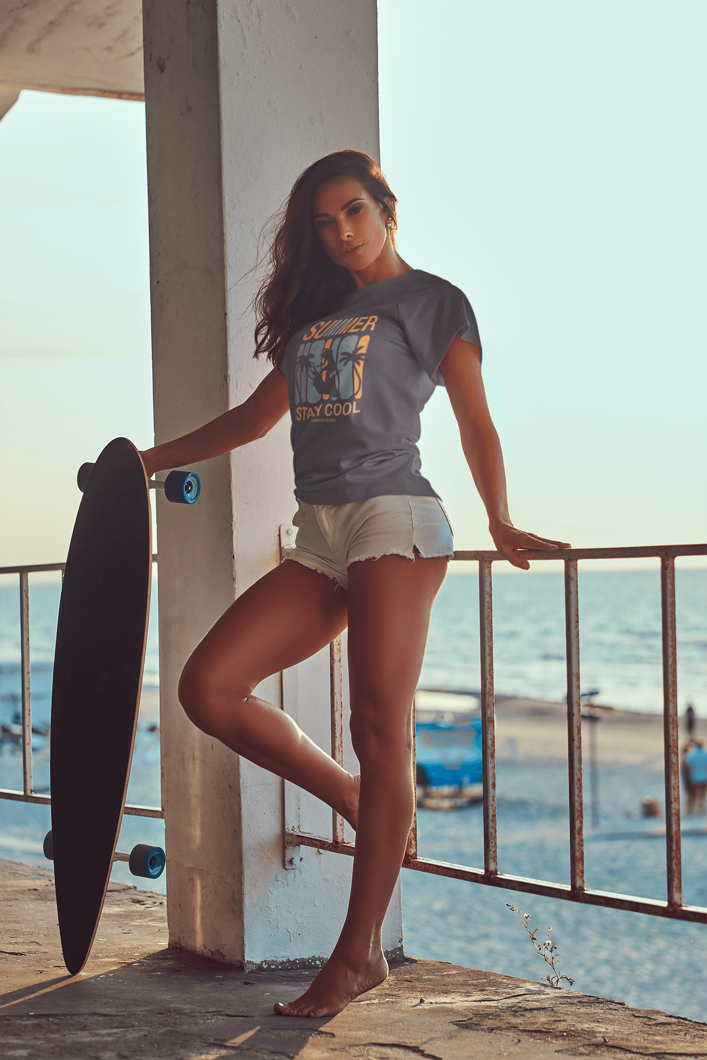 Model wearing Summer Stay Cool T-Shirt with surfer graphic, standing by railing at beach during sunset.