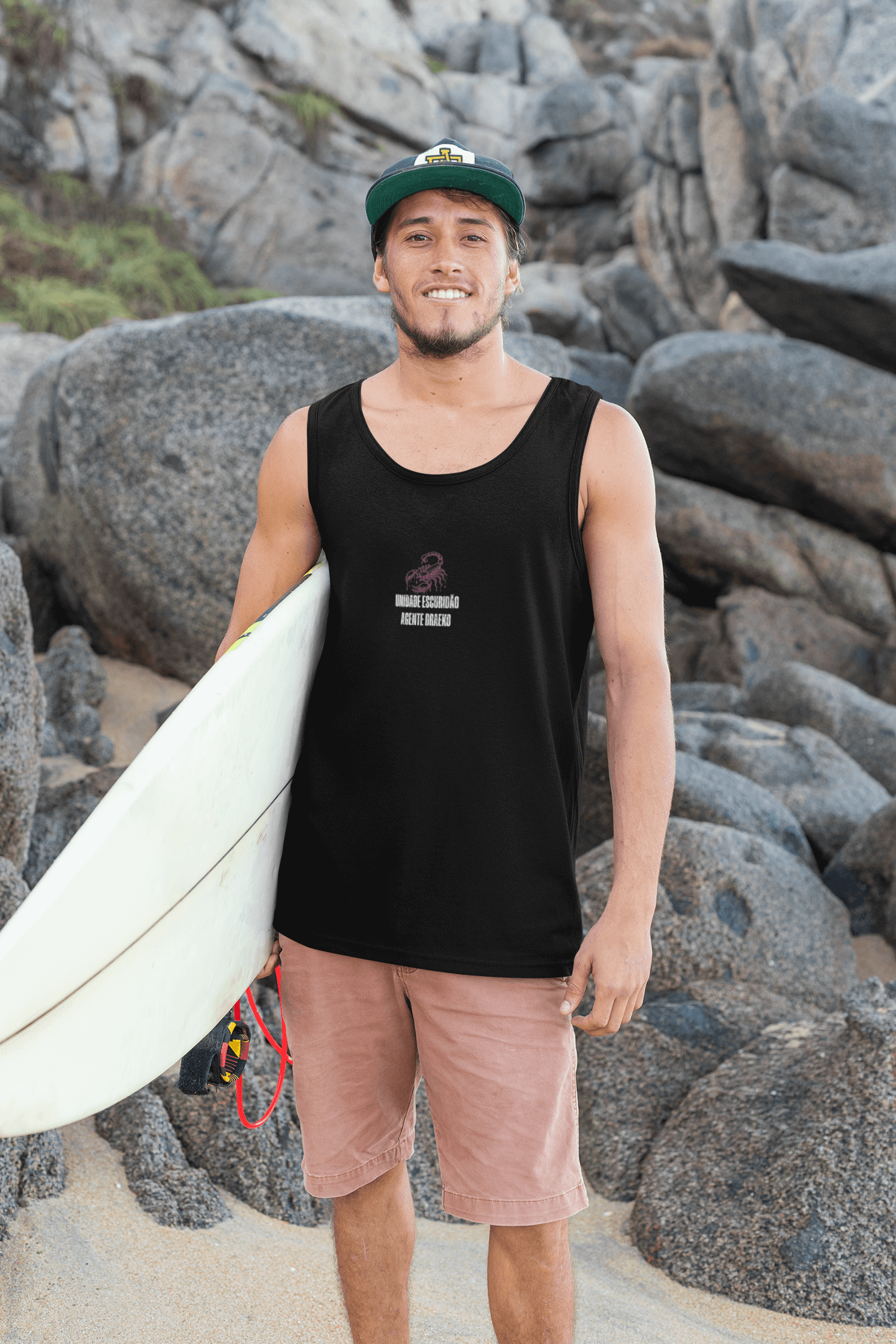 Man wearing Taily Scorpion Graphic Tank Top, holding a surfboard, with rocky background.