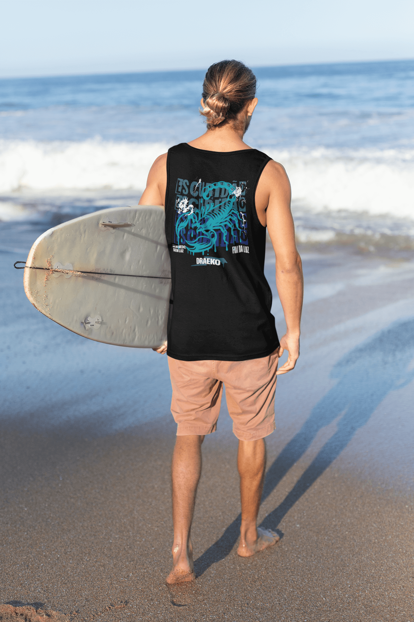 Man wearing Taily Scorpion Graphic Tank Top walking on beach with surfboard, ocean waves in background.