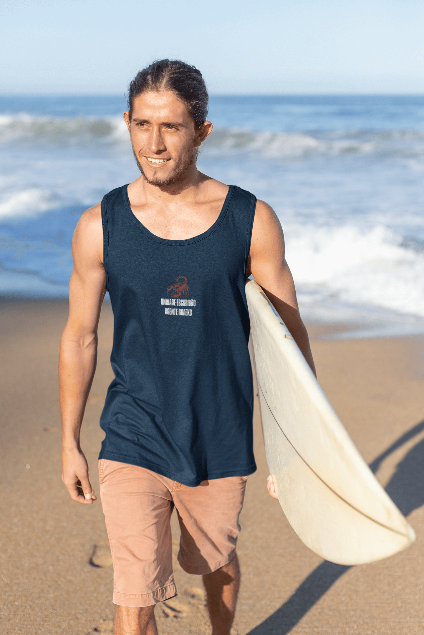 Man walking on the beach wearing the Taily Scorpion Graphic Tank in navy with a surfboard.