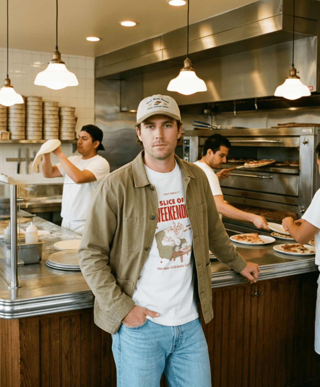 Oversized Pizza T-Shirt – Slice of Weekendin’ (White) worn in a diner setting with pizza chefs in the background.