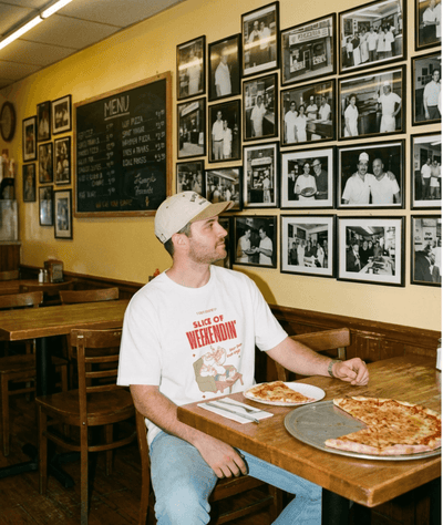 A man wearing an Oversized Pizza T-Shirt – Slice of Weekendin’ (White) sits at a diner table with pizza.