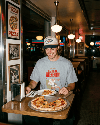 A person wearing an Oversized Pizza T-Shirt – Slice of Weekendin’ (Athletic Heather) enjoys pizza at a diner.