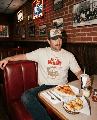 Man wearing Oversized Pizza T-Shirt – Slice of Weekendin’ (Ecru) in diner with pizza and drink.