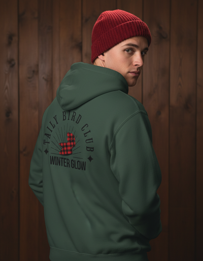 person wearing Taily Bird Club Winter Glow Hoodie in Alpine Green with red beanie against wooden background