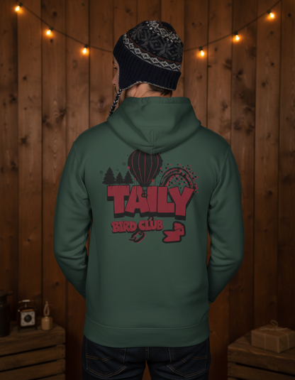 Taily Bird Parkland Hoodie – Alpine Green featuring a colorful graphic on the back in a cozy wooden setting.