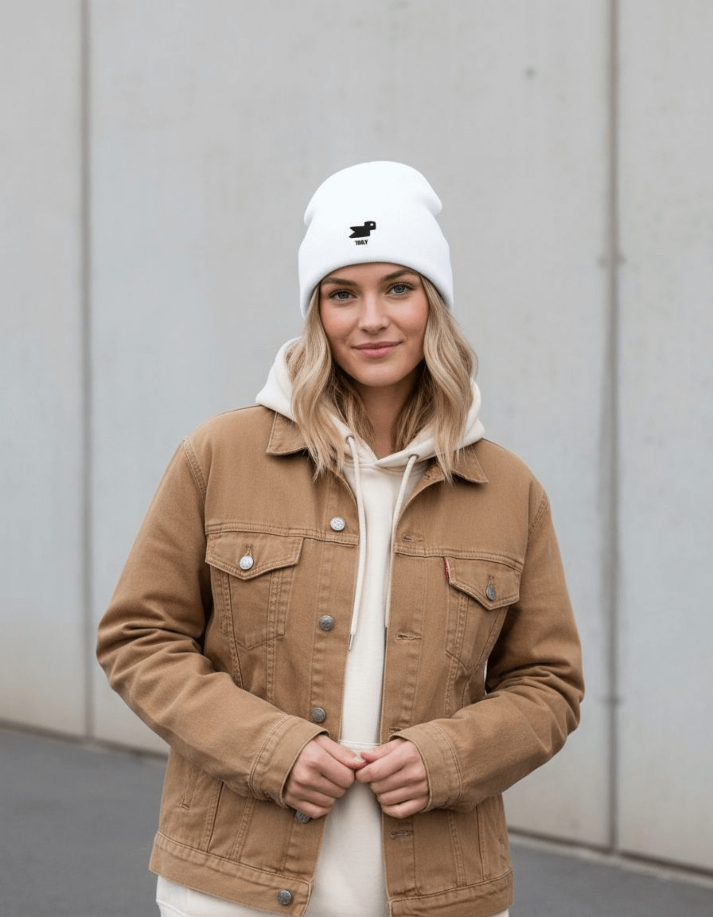 Model wearing Taily Bird Knit Beanie in white with minimal embroidery, paired with a brown jacket.