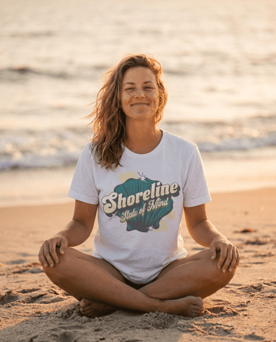 Woman meditating on the beach wearing a Shoreline State of Mind Tee – White with seashell graphic.