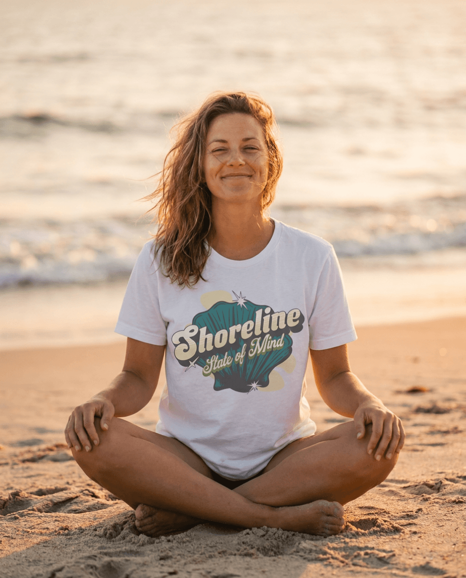 Woman meditating on the beach wearing a Shoreline State of Mind Tee – White with seashell graphic.