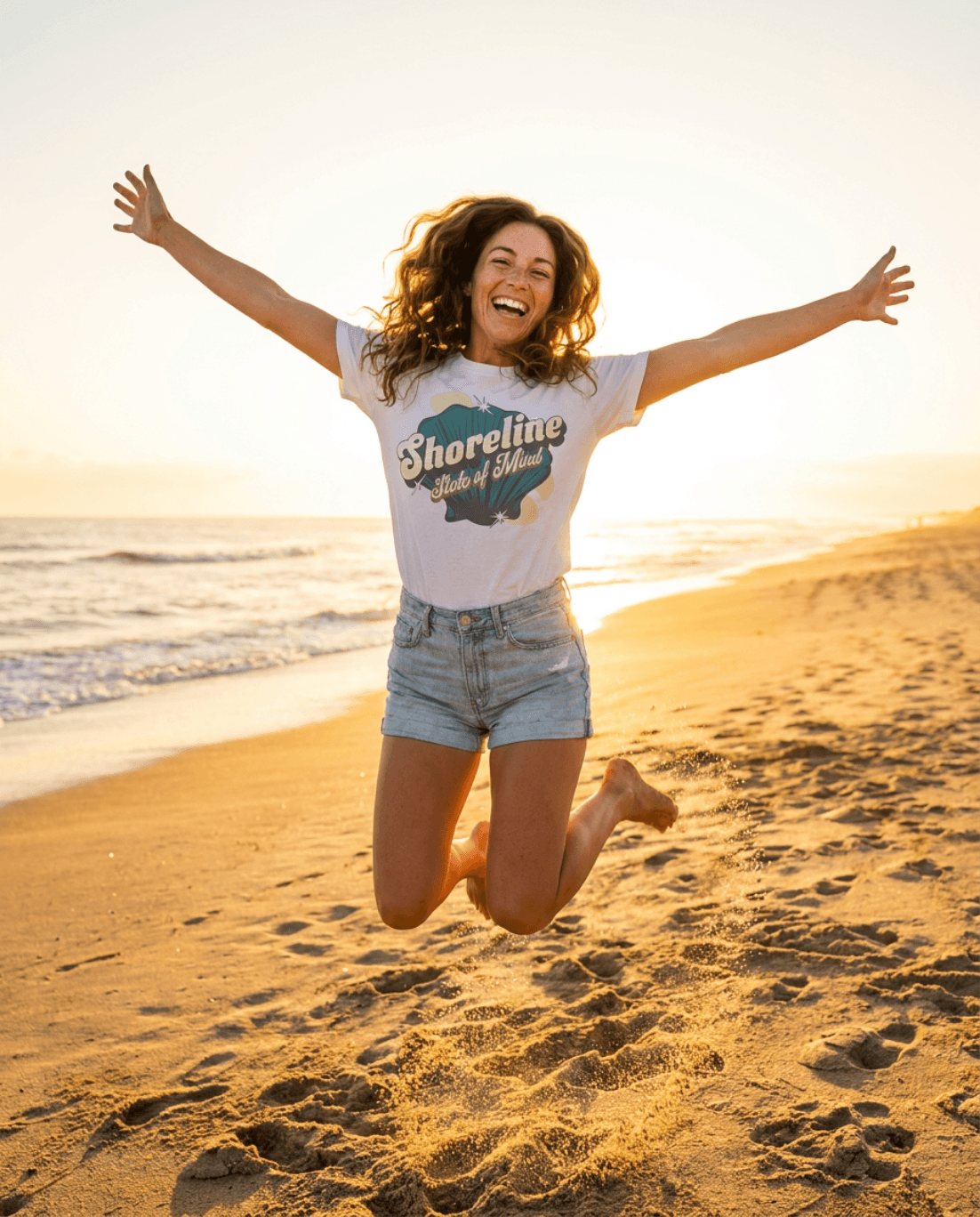 Woman jumping joyfully on the beach wearing Shoreline State of Mind Tee – White at sunset.