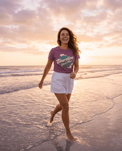 Woman running on the beach at sunset wearing Shoreline State of Mind Tee – Berry with seashell graphic.