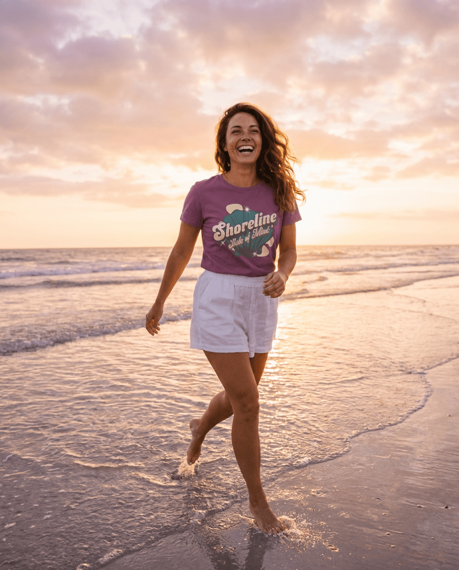 Woman running on the beach at sunset wearing Shoreline State of Mind Tee – Berry with seashell graphic.