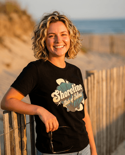 Shoreline State of Mind Tee – Black worn by a woman at the beach, featuring a vintage seashell graphic.