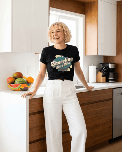 Woman wearing Shoreline State of Mind Tee – Black, smiling in a bright kitchen with a fruit bowl.