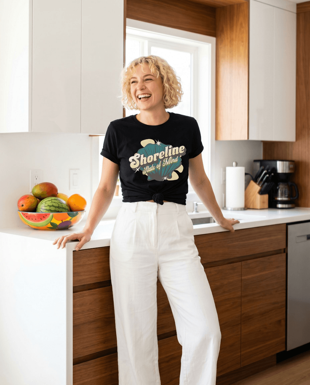 Woman wearing Shoreline State of Mind Tee – Black, smiling in a bright kitchen with a fruit bowl.