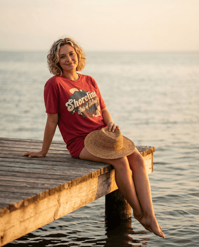Model wearing Shoreline State of Mind Tee – Crimson, sitting on a dock by the water during sunset.