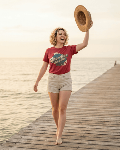 Woman wearing the Shoreline State of Mind Tee – Crimson, walking on a pier and holding a straw hat.