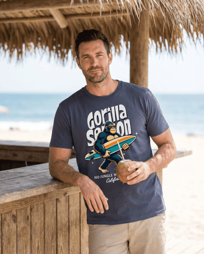 Man wearing Gorilla Season Surf Tee – Navy, holding a coconut, with a beach backdrop.