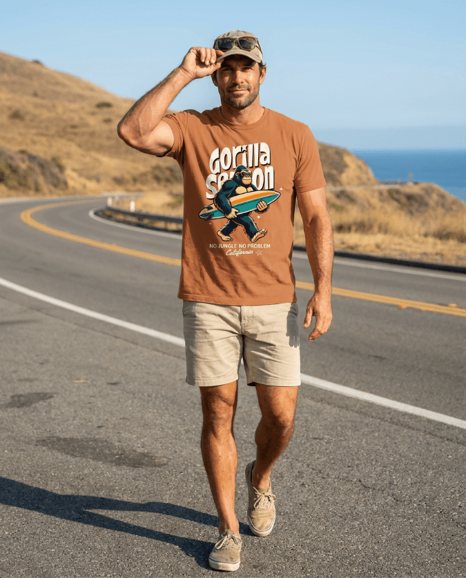 Man walking on a road wearing a Gorilla Season Surf Tee – Yam and shorts against a coastal background.