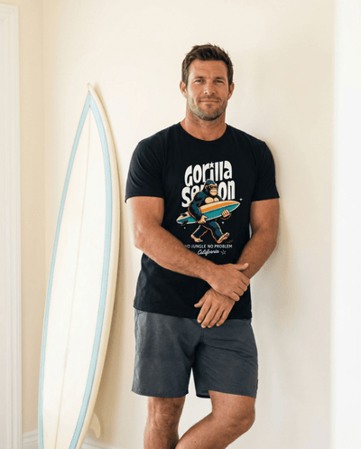 Man wearing Gorilla Season Surf Tee – Black standing next to surfboard, showcasing California surf graphic.