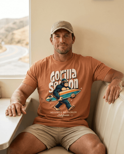 A man wearing a Gorilla Season Surf Tee – Yam, featuring a laid-back gorilla graphic, sitting indoors.